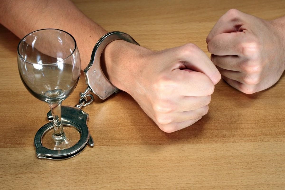 Руки на фоне стола. Одна рука пристёгнута наручниками к пустому бокалу